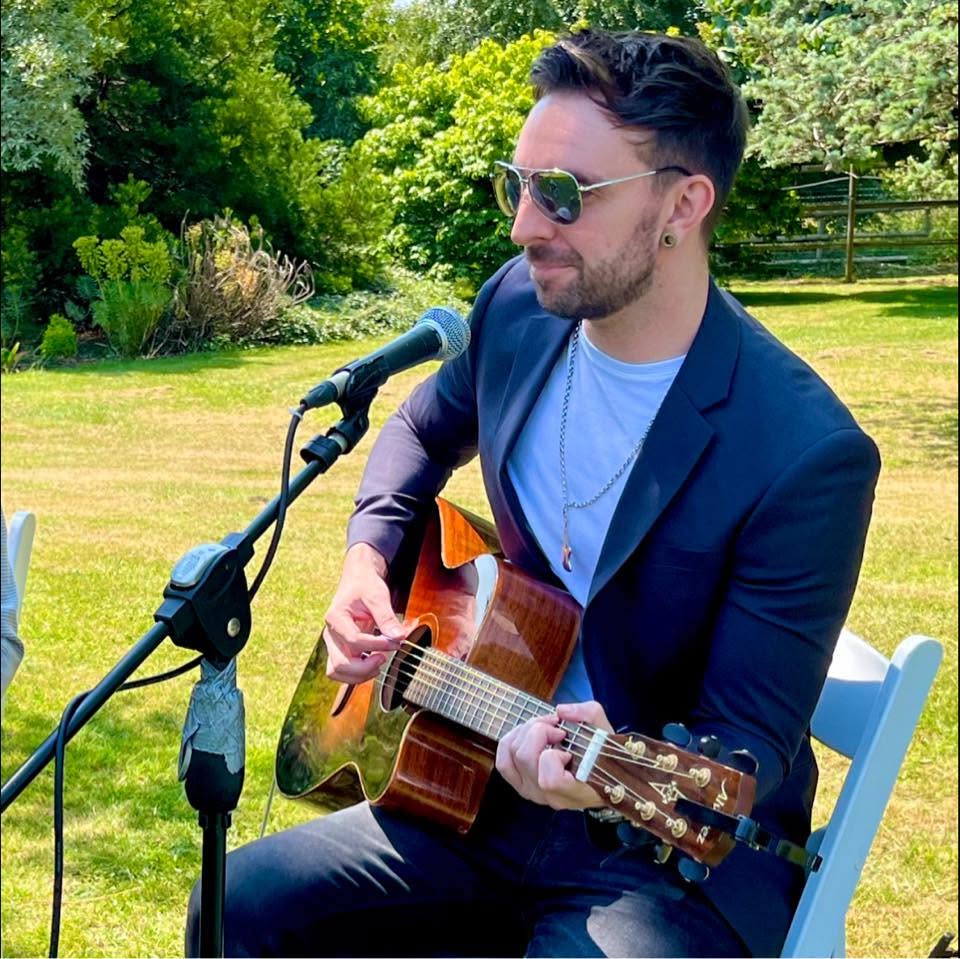 Rob Galley in a navy blazer and sunglasses, playing an acoustic guitar at a microphone on a sunny lawn